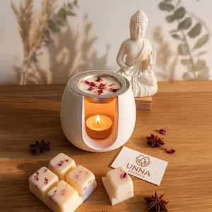 Quemador de aceites esenciales de cerámica blanca con vela tealight encendida, acompañado de pastillas de cera de soja con pétalos de rosa, anís estrellado y tarjeta de UNNA Essence sobre superficie de madera.