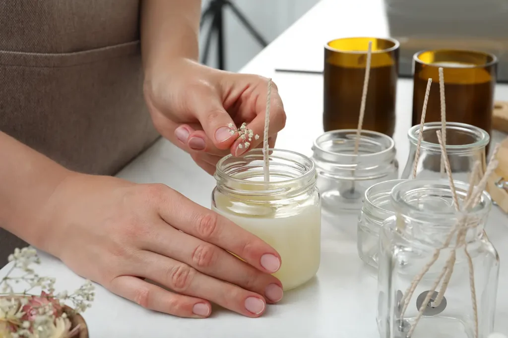 Primer plano de unas manos artesanales colocando delicadas flores de gipsófila seca dentro de un frasco de vidrio lleno de cera derretida para hacer una vela natural. En primer plano, otros frascos con mechas preparadas y vasos ámbar de fondo, en un taller de fabricación de velas.