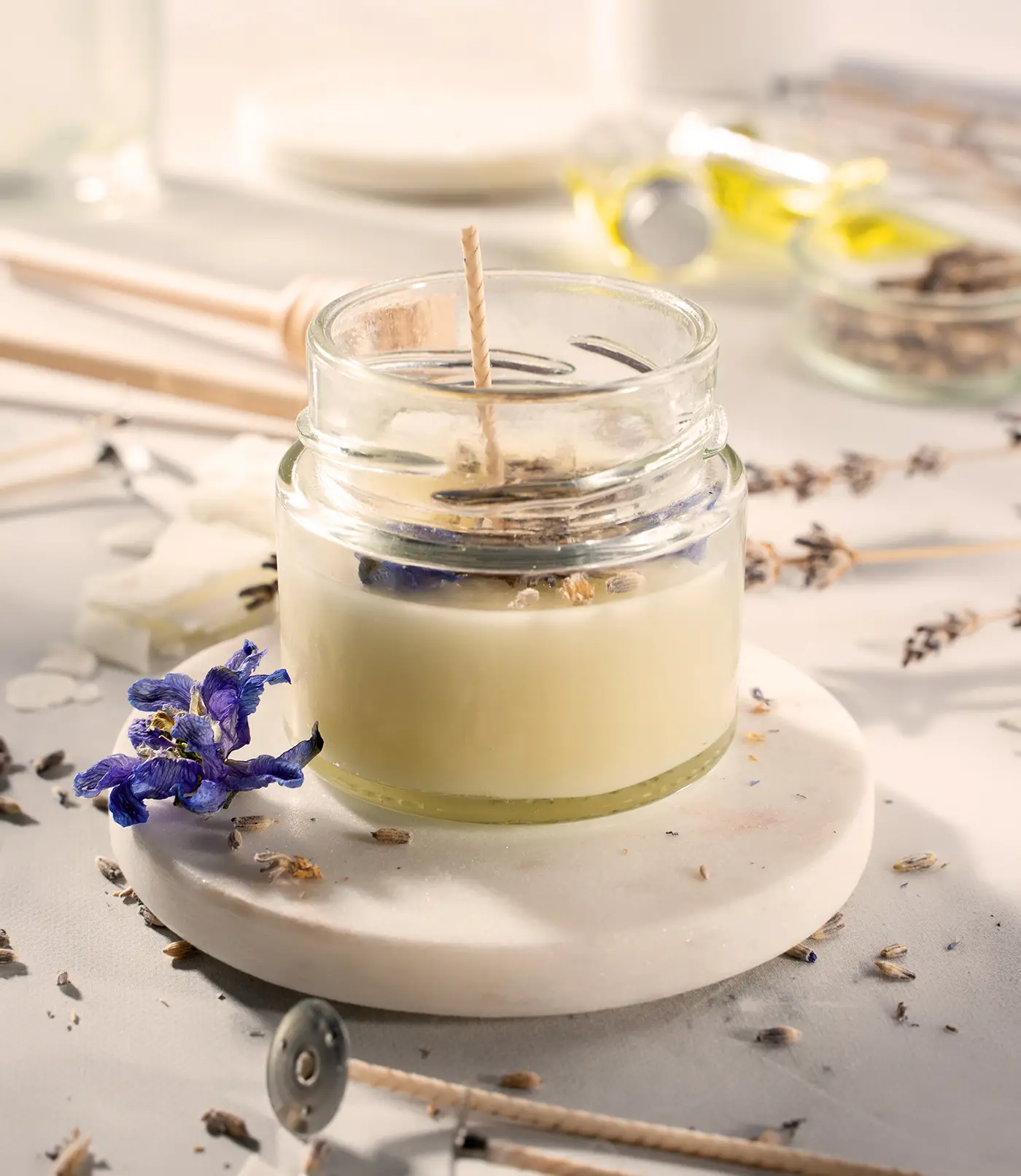 Vela artesanal de cera de soja con flores de lavanda y aroma relajante en un tarro de cristal transparente sobre una base de mármol redonda. Al lado de la vela hay una rama de flores de lavanda y una caja de cerillas. La vela está encendida.