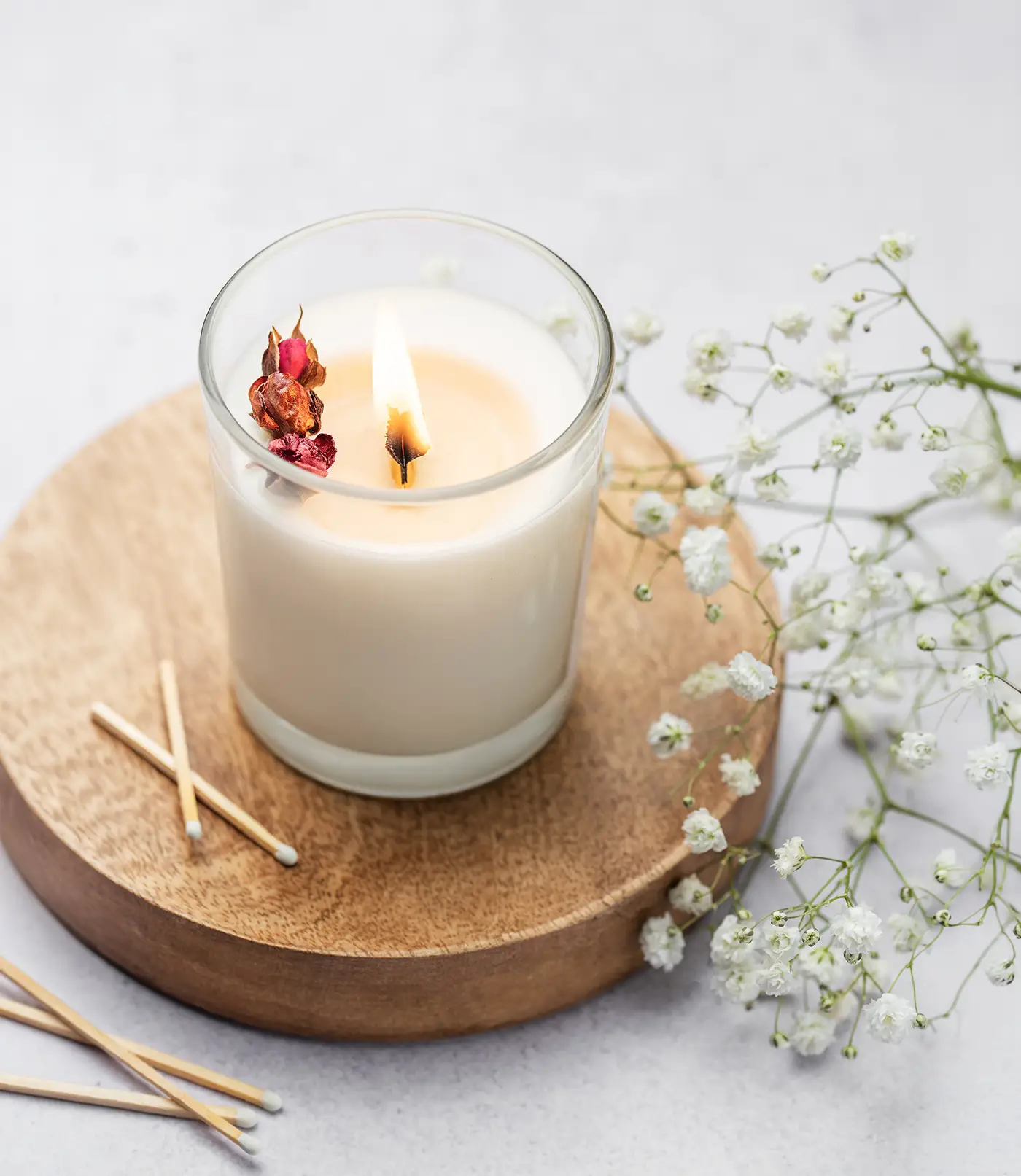Vela artesanal de cera de soja con pétalos de rosa y aroma floral en un tarro de cristal transparente sobre una base de madera redonda. Al lado de la vela hay una rama de flores blancas y una caja de cerillas. La vela está encendida.